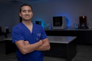 Man in blue scrubs stands confidently in a dimly lit lab room with arms crossed, smiling at the camera.