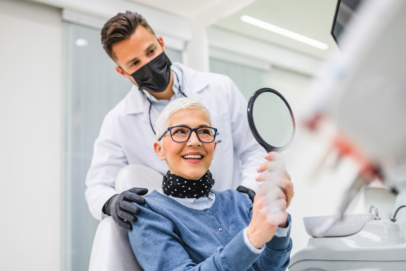an older patient smiling at herself through a hand held mirror she's holding in front of herself with the doctor smiling over her with his dental mask on because she is a candidate for dental implants.